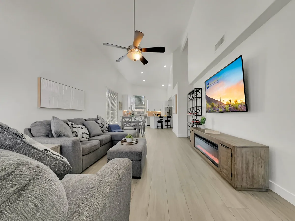 This image showcases a spacious gray sectional sofa arranged for conversation and relaxation, a modern TV unit with decorative wall art, and a dining area visible in the background. Floor-to-ceiling windows allow natural light to wash over the space, hinting at scenic views beyond.