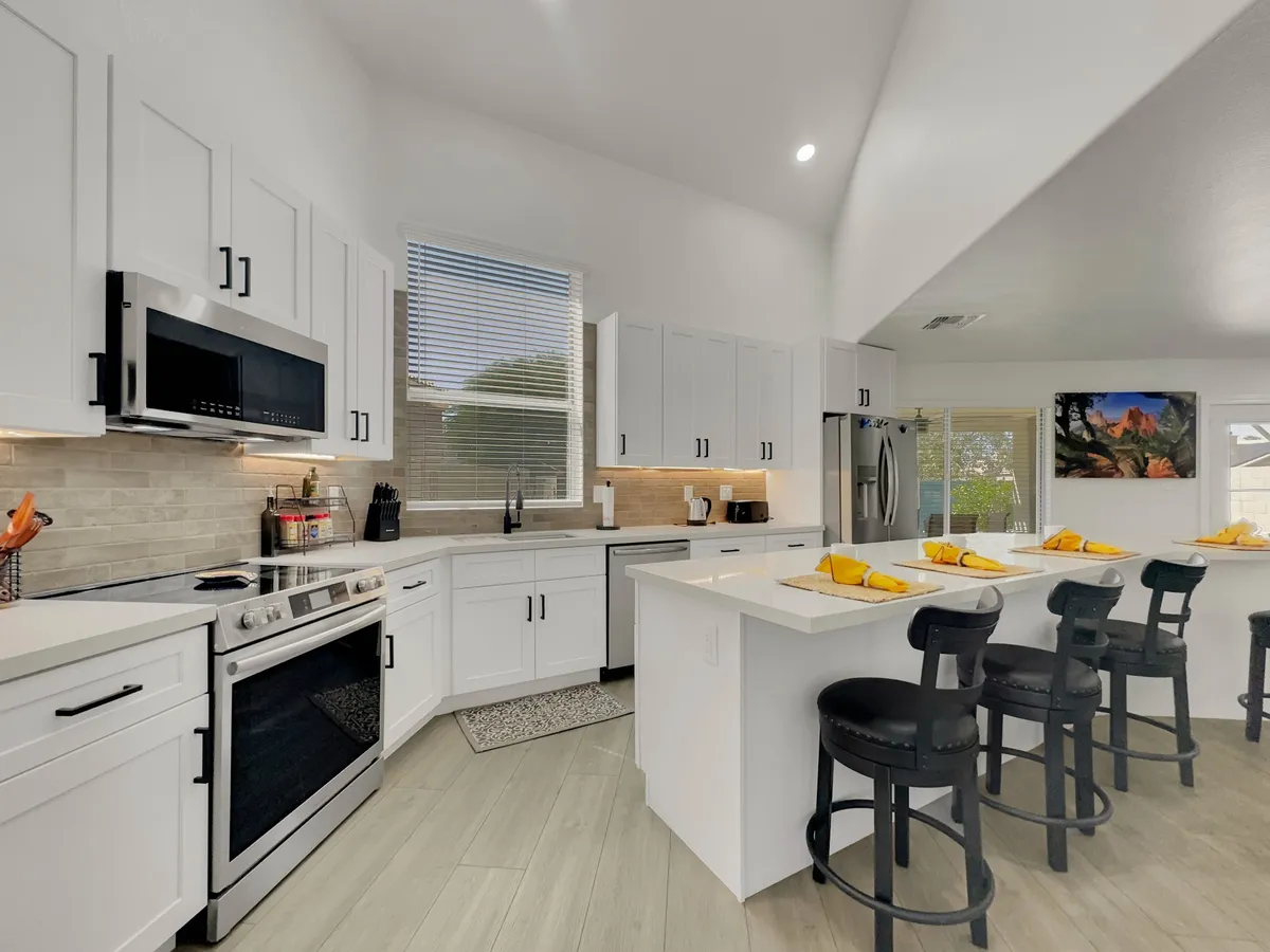 A bright, spacious kitchen featuring a large island with bar stools, gleaming stainless steel appliances, white cabinetry, and a large window that fills the space with natural light. The area is designed for both meal prep and social gatherings, with a layout that encourages interaction.