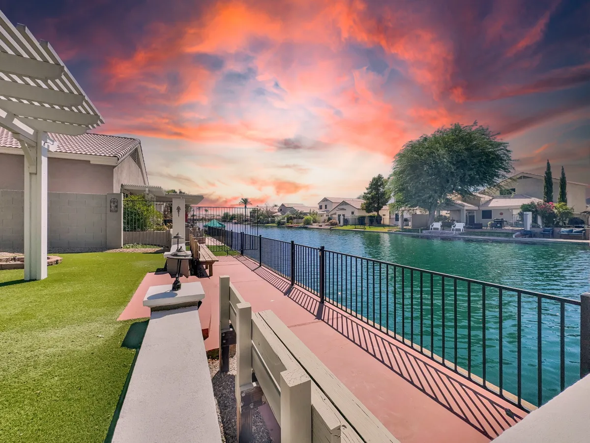 A scenic waterfront view from the yard, with a lush green lawn leading to a fenced walkway along the water. The sky is painted in sunset hues, and the tranquil water reflects the colors beautifully. The area looks perfect for evening strolls, relaxation, and outdoor activities.