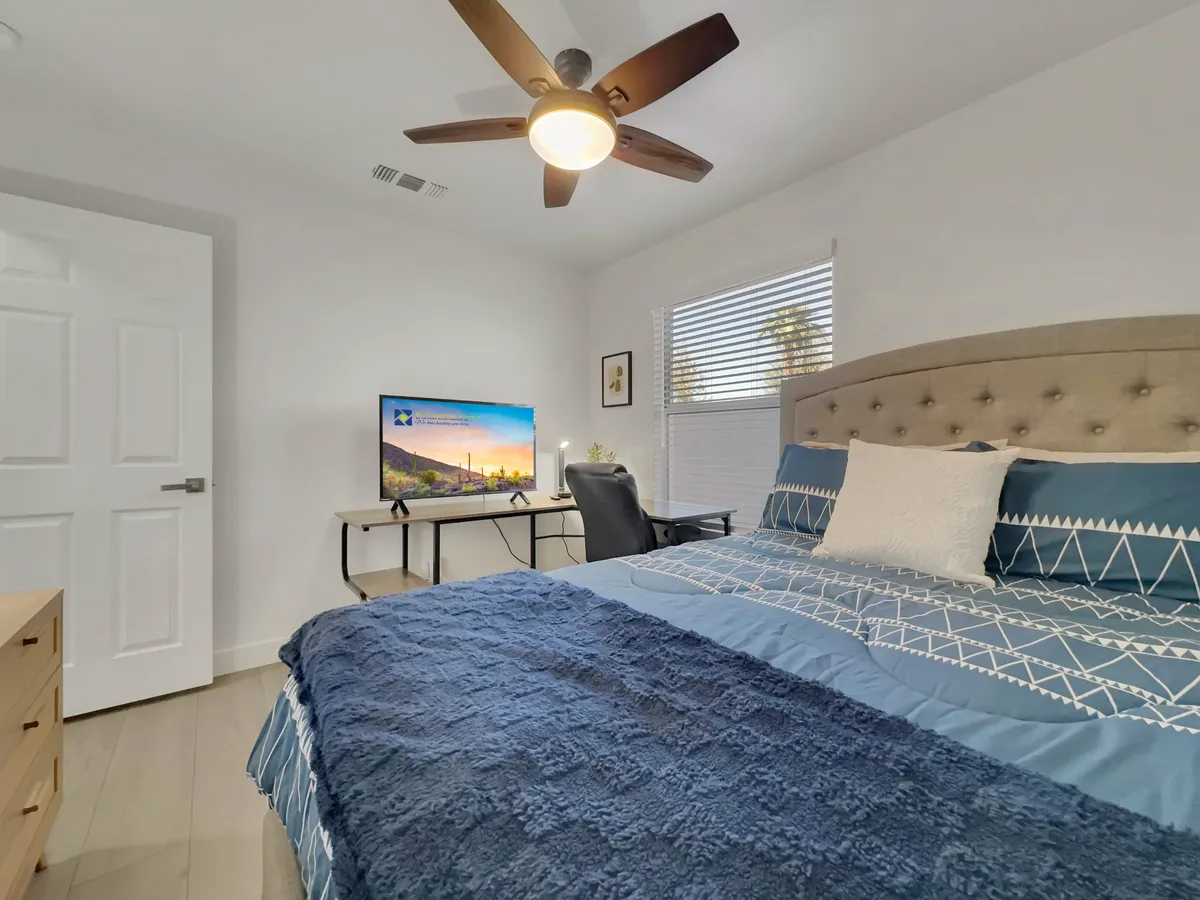 A comfortable bedroom setup doubling as an office, with a bed adorned in decorative pillows, a desk with a monitor for remote work, and a ceiling fan for comfort. The space is both functional and cozy, ideal for guests or productive work-from-home days.
