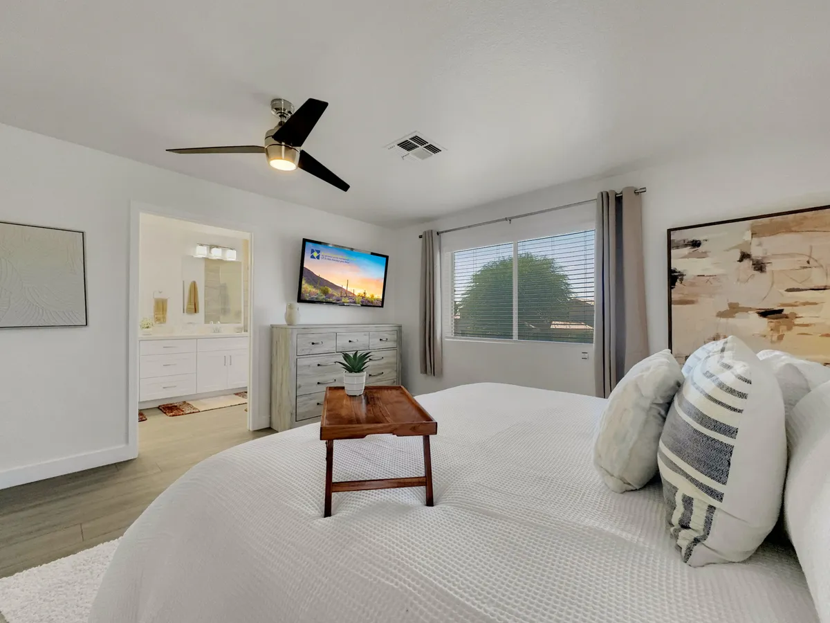 A serene bedroom featuring a king-sized bed with textured white bedding, a wooden tray for morning coffee, a wall-mounted television with a scenic background, large windows allowing abundant natural light, and abstract artwork. The atmosphere is calm and inviting, ideal for restful nights and peaceful mornings.