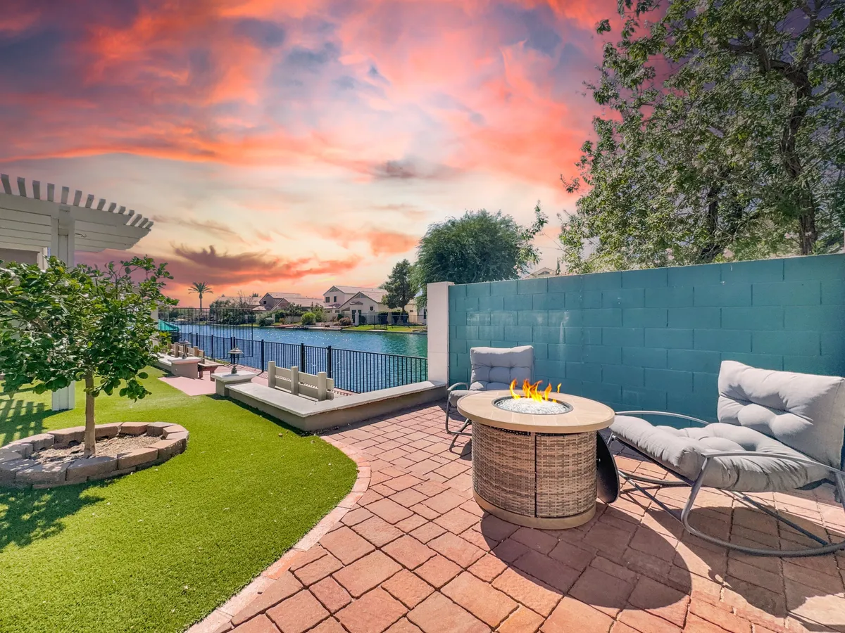 A tranquil backyard scene featuring a glowing fire pit surrounded by comfortable lounge chairs. The water view dominates the background, and the sky is painted with vibrant sunset colors. The setup suggests a perfect place for family or friends to gather, relax, and enjoy evening conversations as the sun sets over the water.