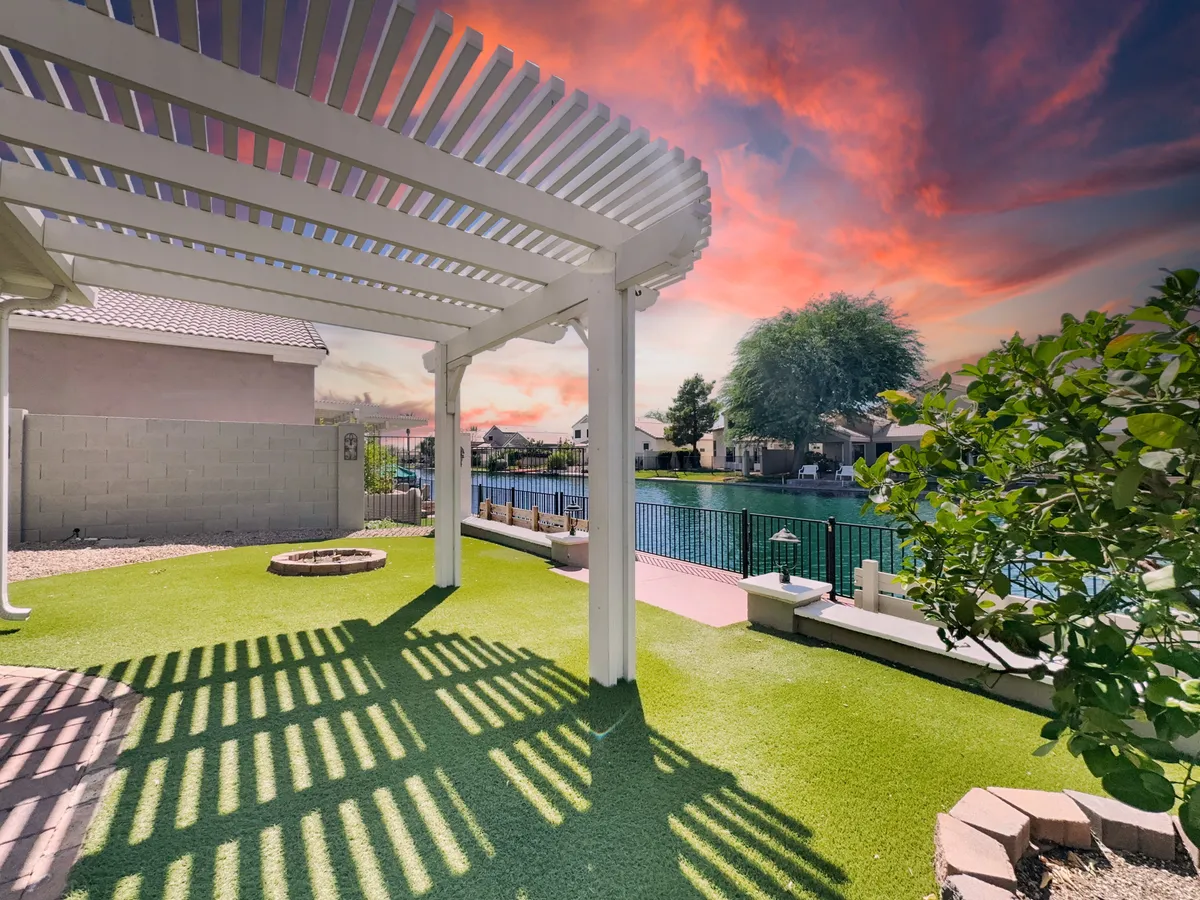 A stylish pergola shades an artificial grass area with a modern fire pit and lounge seating, set beside a peaceful waterfront. The view captures the sunset, making it ideal for entertaining or relaxing outdoors.