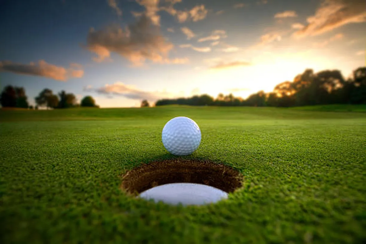 A serene golf course scene featuring a close-up of a golf ball near the hole on a lush green, bathed in the golden light of sunset. The expansive green space and the sense of leisure reinforce the leisurely lifestyle available nearby.