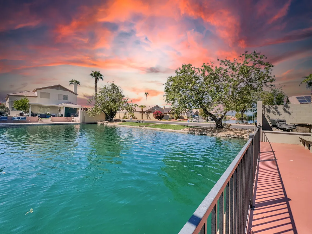 This image captures a tranquil waterfront at sunset, with calm water, lush green trees, colorful sky, and residential homes lining the shore. The inviting natural setting hints at peaceful walks and relaxing moments by the water.