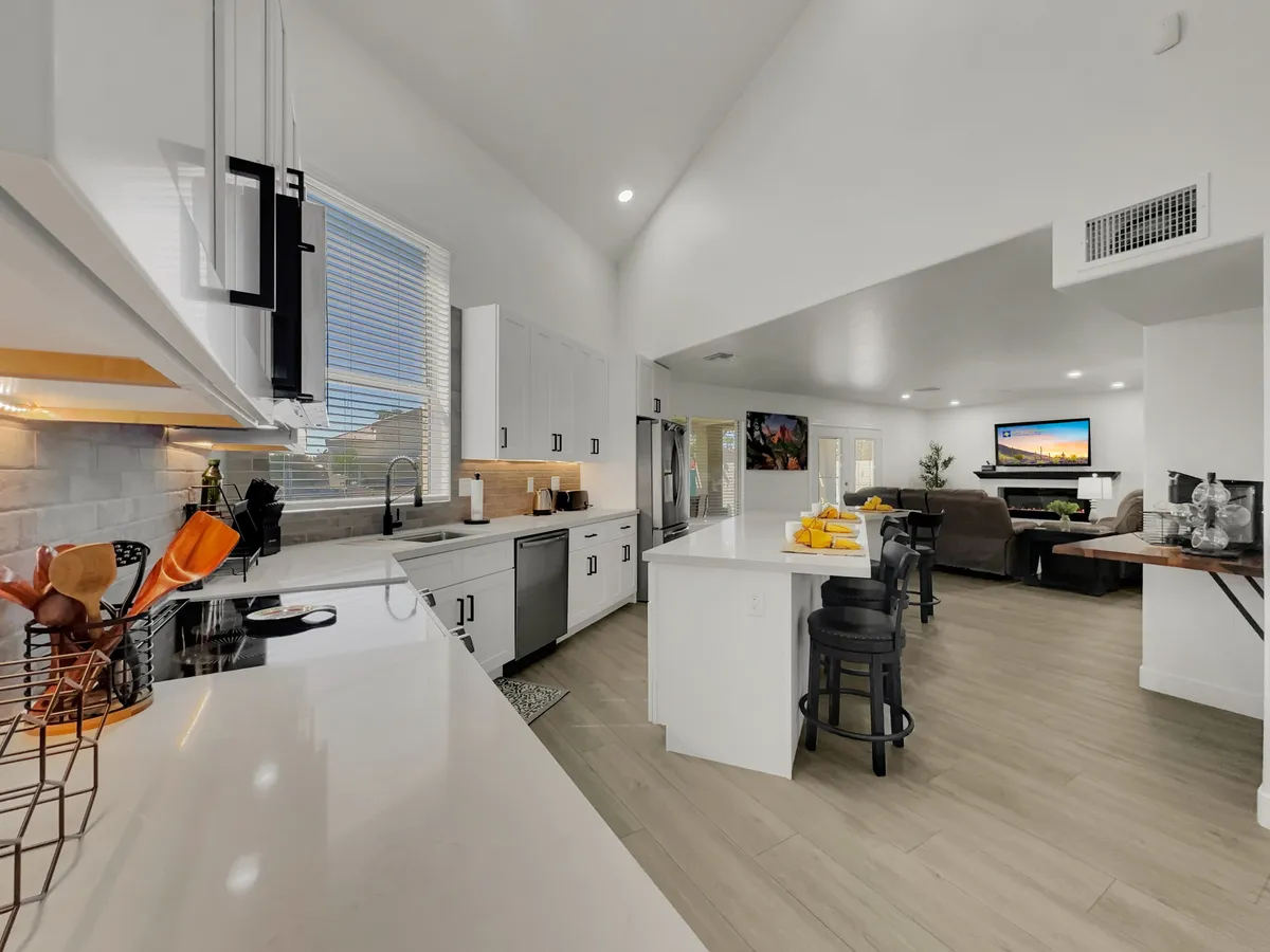 An inviting kitchen island with a colorful fruit display is the focal point, surrounded by sleek modern appliances and open sightlines to the living area. The design is airy and welcoming, emphasizing both culinary creativity and social connection.